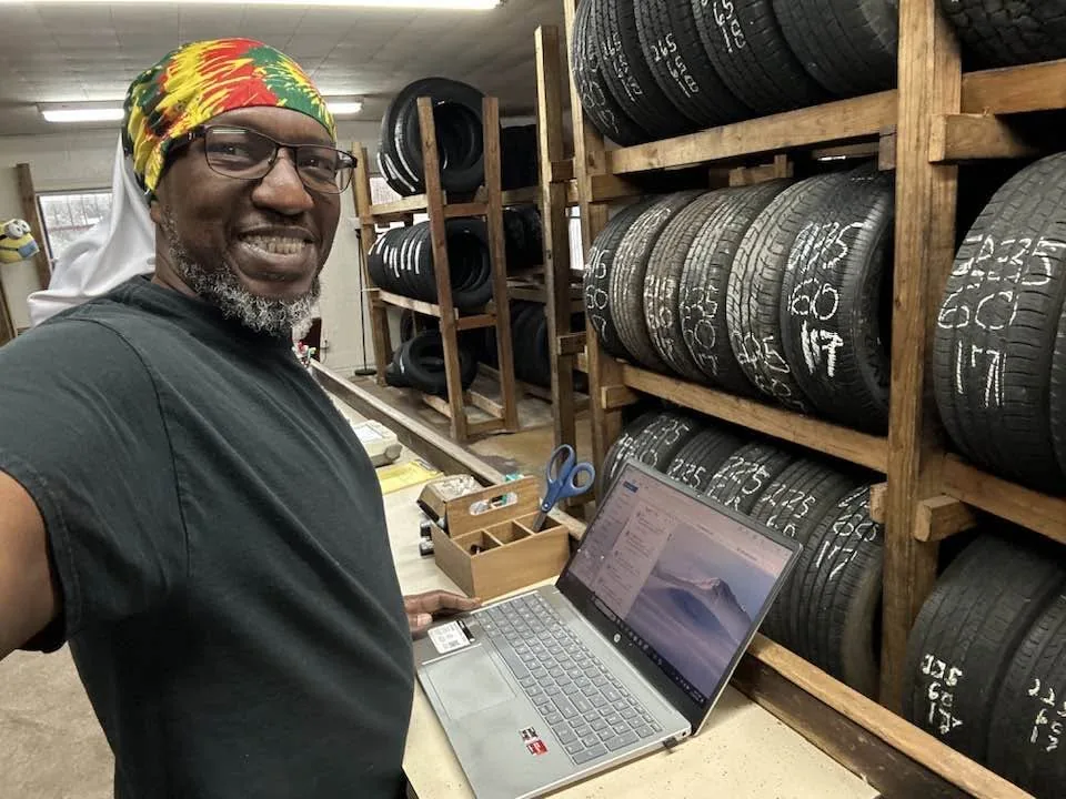 Bobby Cato smiling inside the shop, in front of wooden racks full of used tires with their sizes written on the sidewalls.